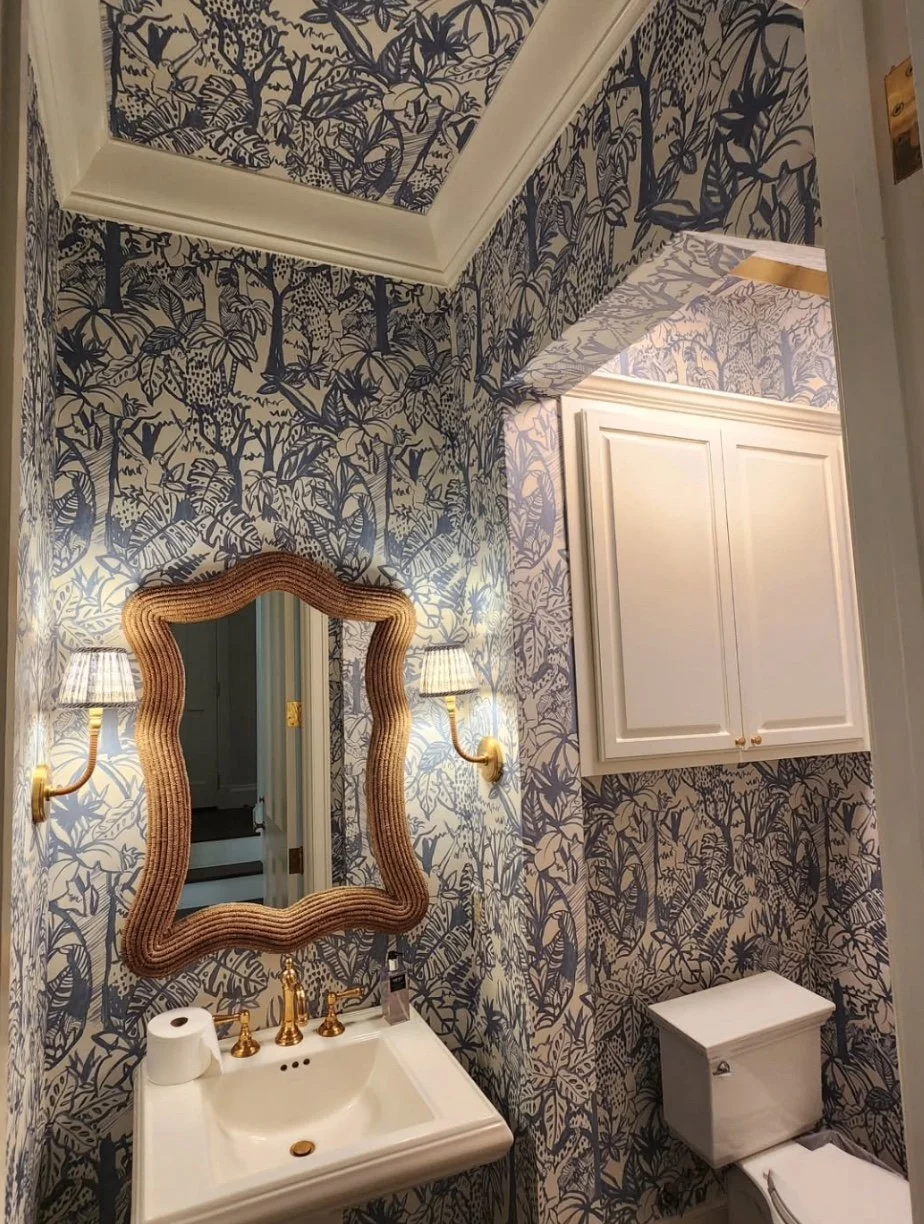A bathroom with floral-patterned wallpaper, a white sink with gold fixtures, a framed mirror, wall-mounted lamps, a white cabinet above the toilet, and a toilet with a tank.