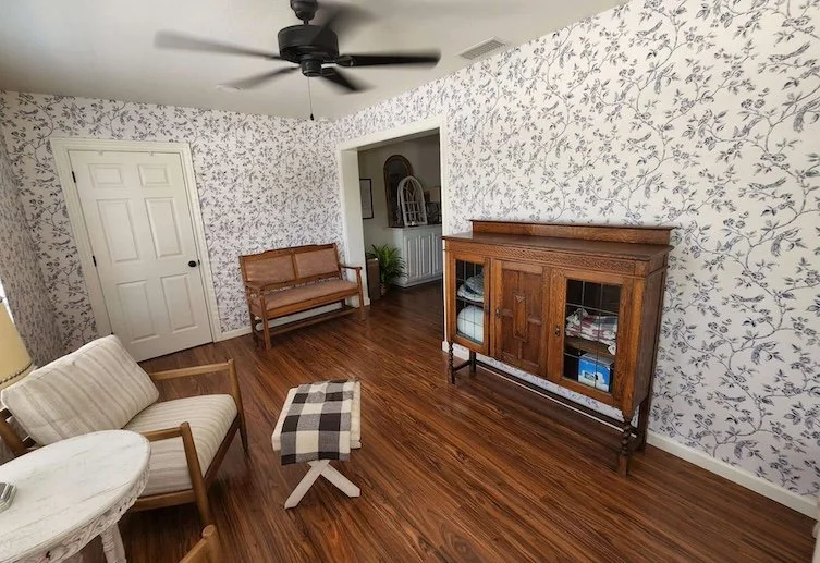 Living room with wood flooring, floral wallpaper, a white door, ceiling fan, wooden bench, small table with striped cushion, wooden cabinet with glass doors, and potted plant.