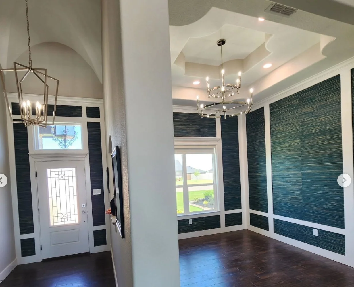 The image shows a modern, well-lit reception area of a house with a decorative entry door. The space features high ceilings, a chandelier, and stylish wall paneling in blue and white. Large windows allow natural light to brighten the room, and dark h