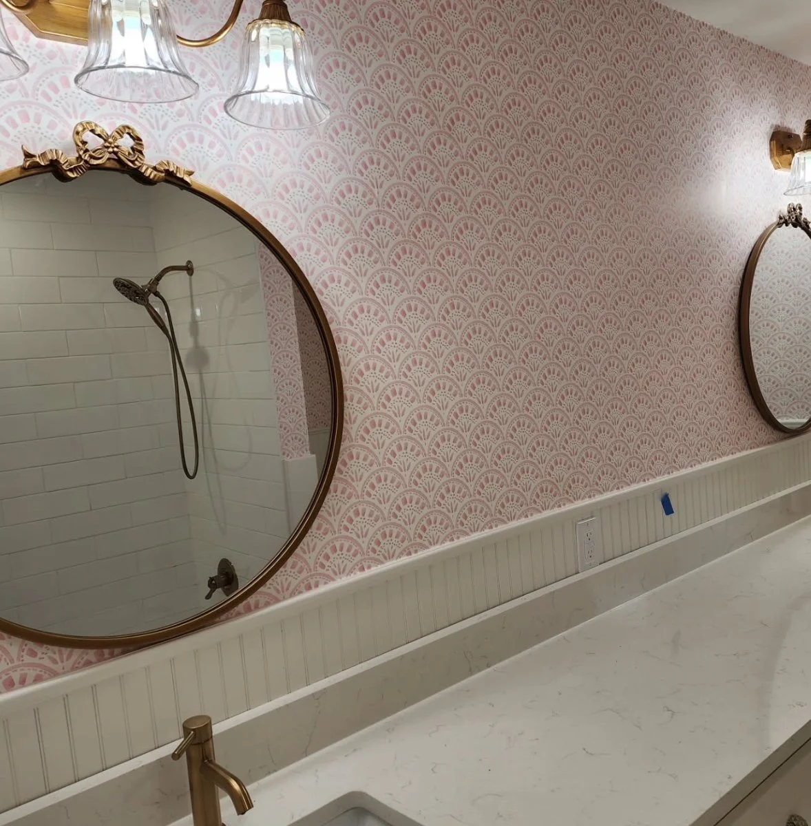 Bathroom with pink patterned wallpaper, oval mirror, white beadboard wainscoting, marble countertop, and wall-mounted light fixtures.