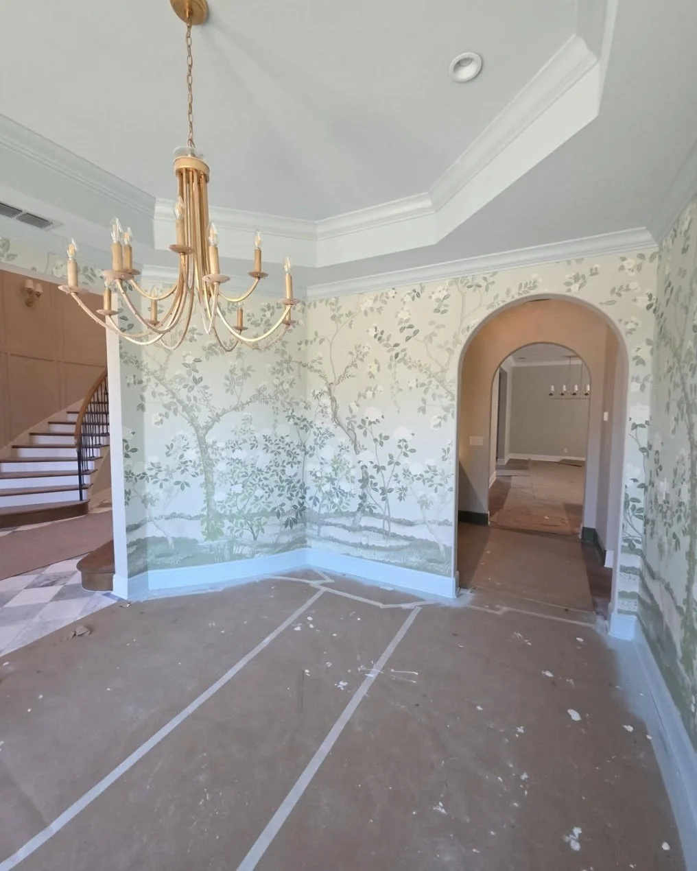 Room with painted wallpaper depicting trees and branches, chandelier hanging from the ceiling, and an arched doorway leading to another room. The floor appears to be under renovation.