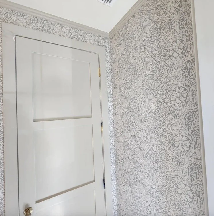 A closed white interior door with gold hinges and a rounded brass doorknob, set in a room with floral patterned wallpaper in light gray and white, with an air vent visible on the ceiling.