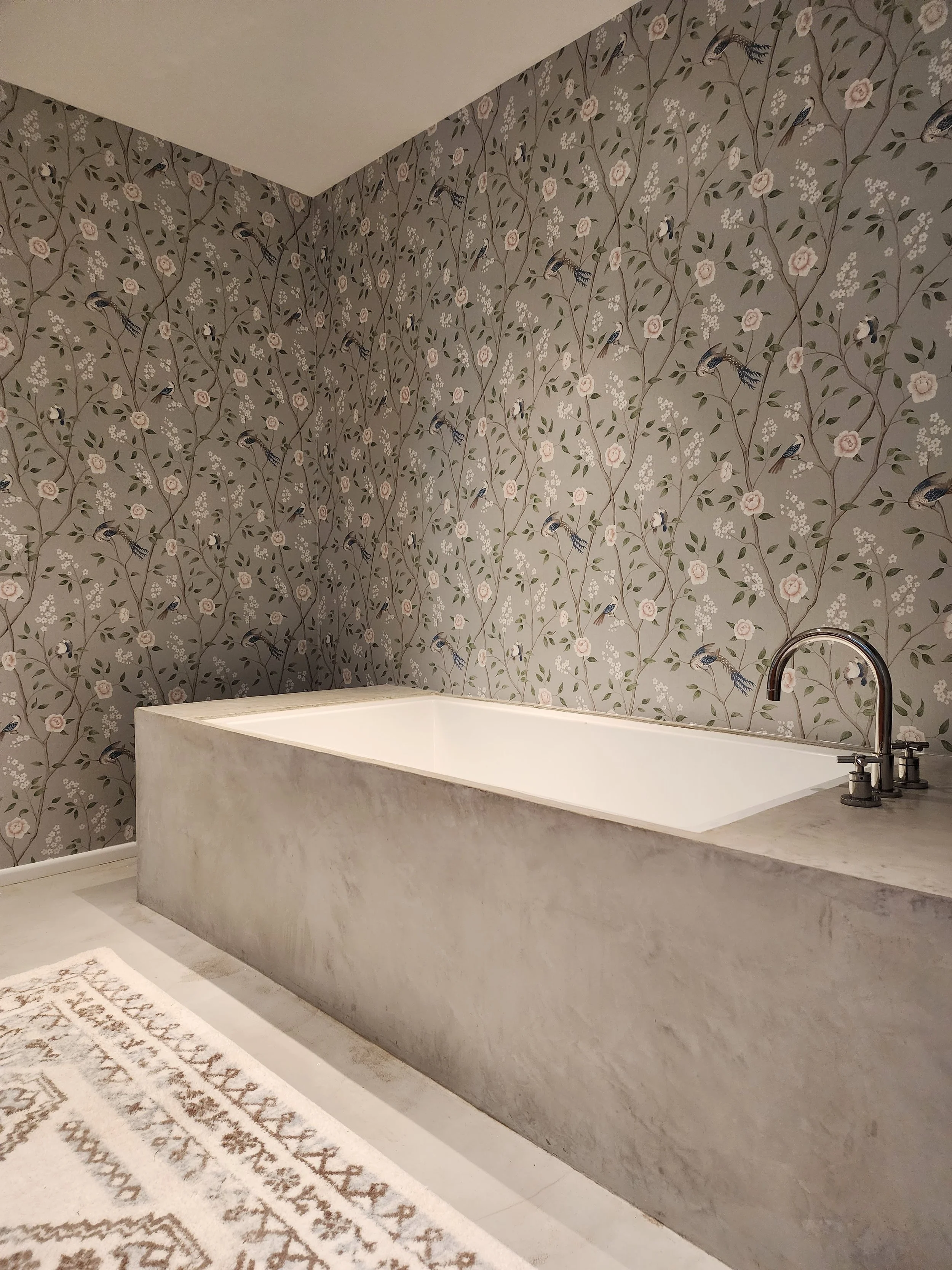 Bathroom with a large bathtub, grey marble exterior, floral wallpaper with pink roses and blue birds, white floor, and a patterned rug.