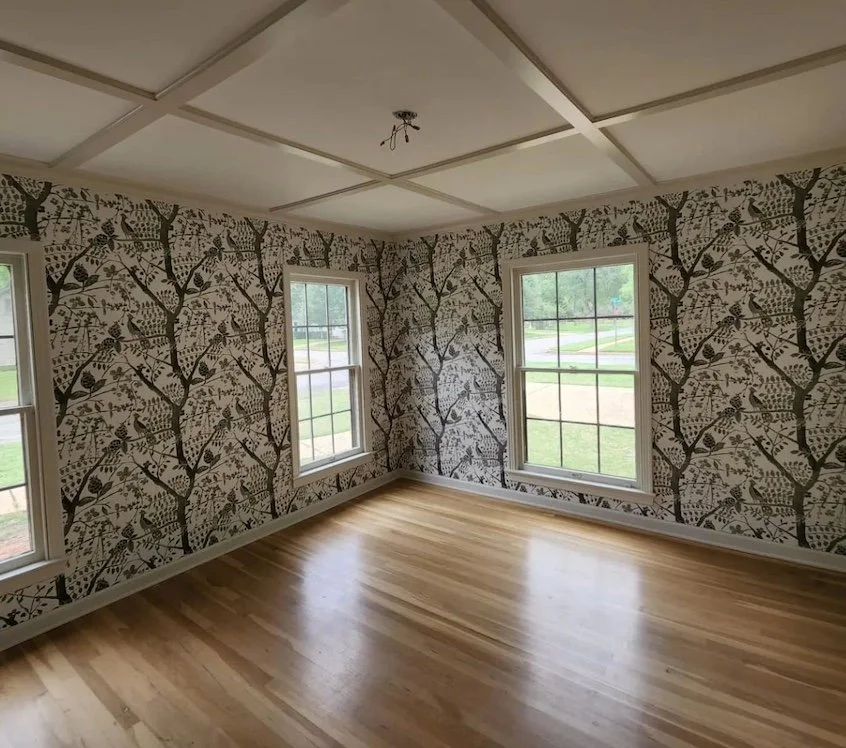 Empty room with three windows, patterned wallpaper with tree and bird design, and light-colored wooden floor.