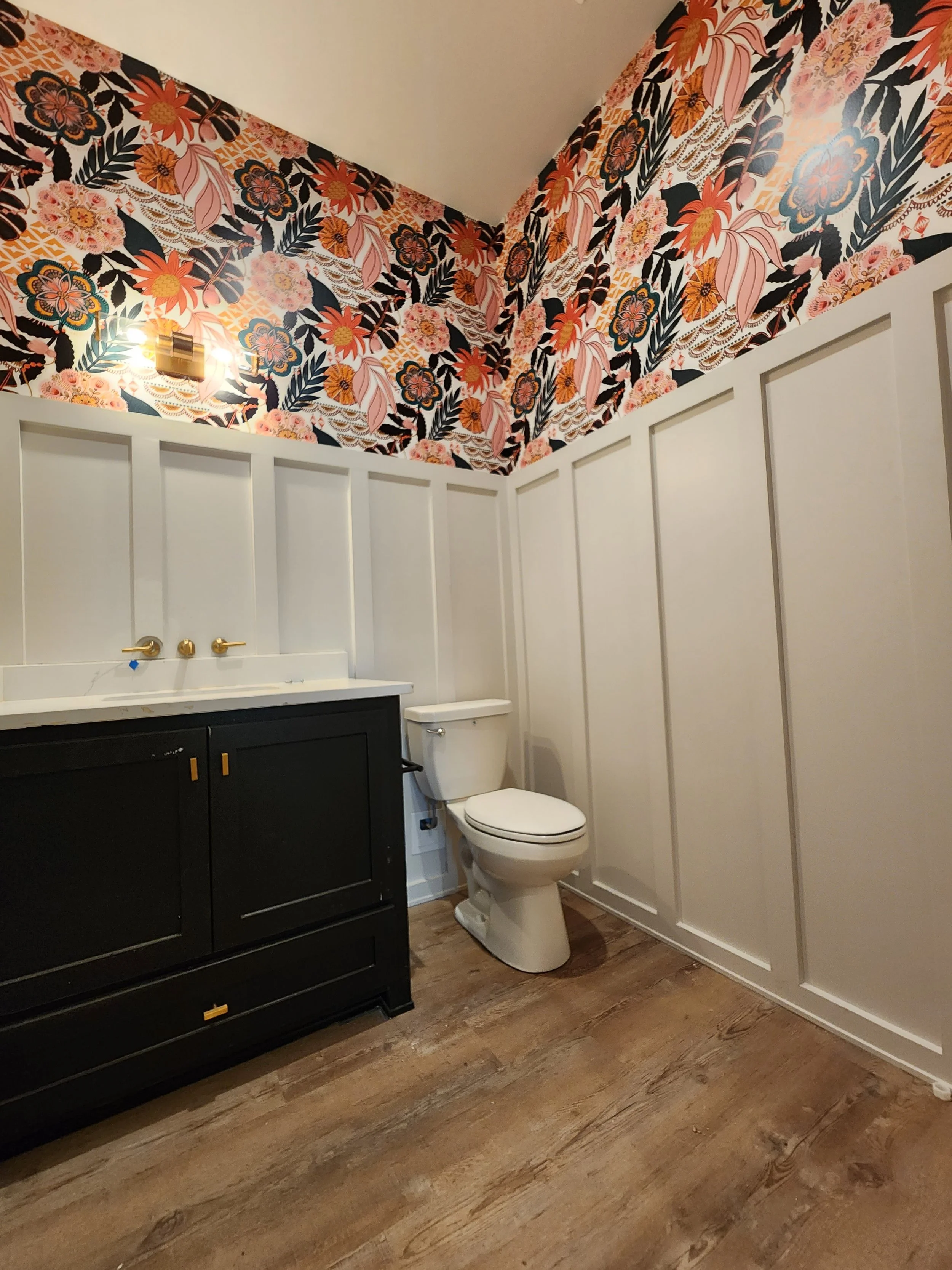 A bathroom with floral wallpaper, a vanity with a white countertop, a toilet, and wood flooring.