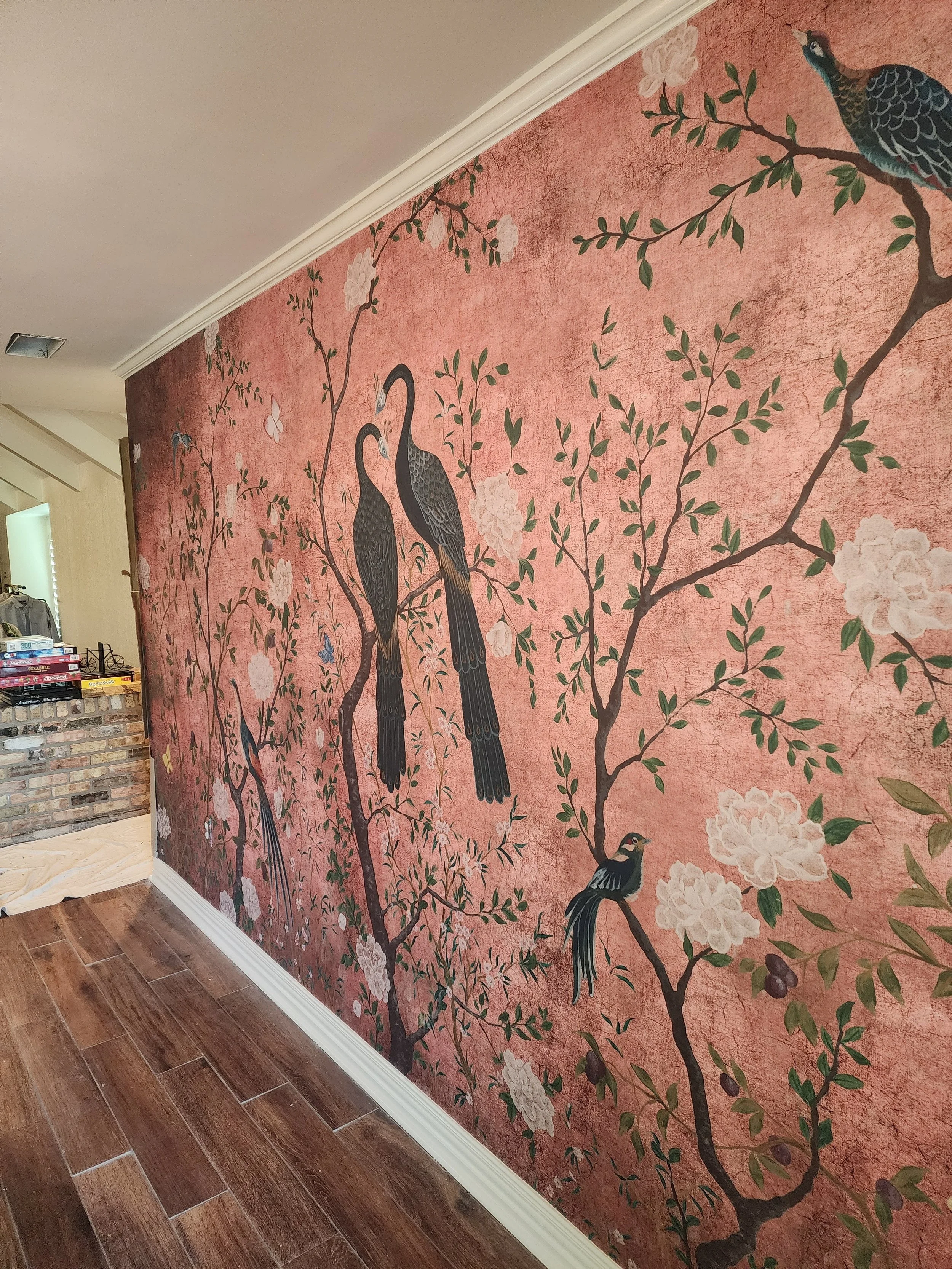 Decorative wall mural featuring branches with white flowers and green leaves, with various black and colored birds perched on the branches, painted on a pink textured background.