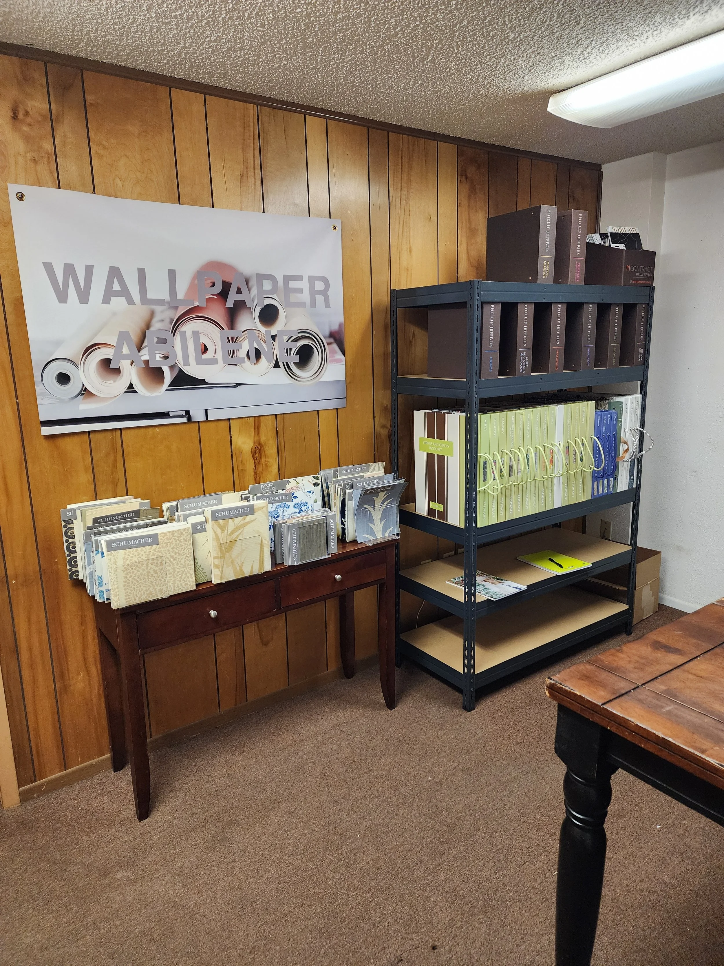 Interior of a room with wood-paneled wall, a poster reading 'WALLPAPER AVAILABLE,' a dark bookshelf filled with binders and boxes, a small wooden table with sample wallpaper books, and part of a wooden desk.