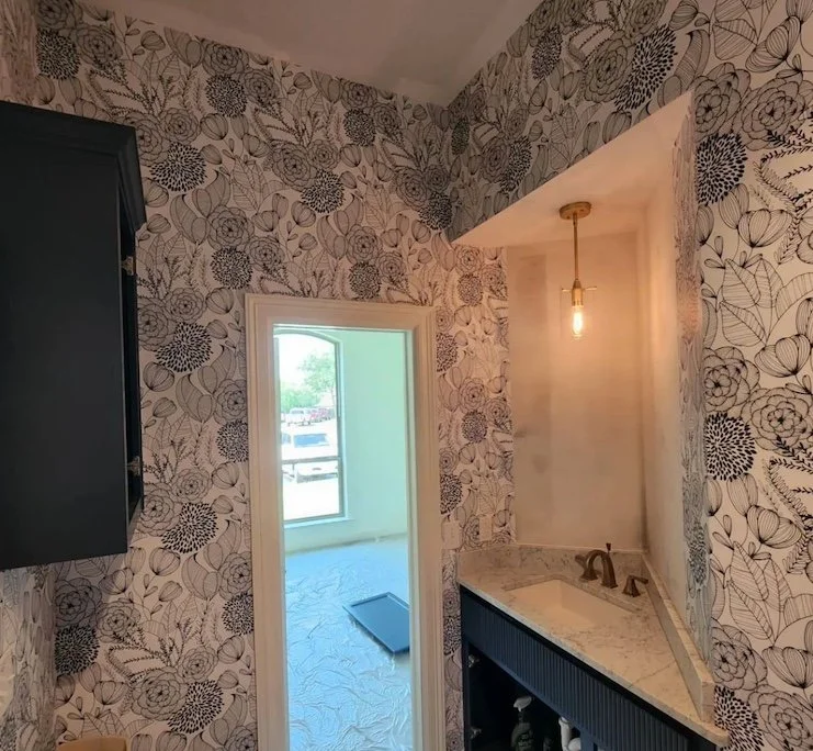 A small room with floral patterned wallpaper, a black cabinet on the left, a doorway leading to a bright room with a window, and a marble countertop with a brass faucet beneath a hanging light fixture. There is also a dark-colored cabinet below the c