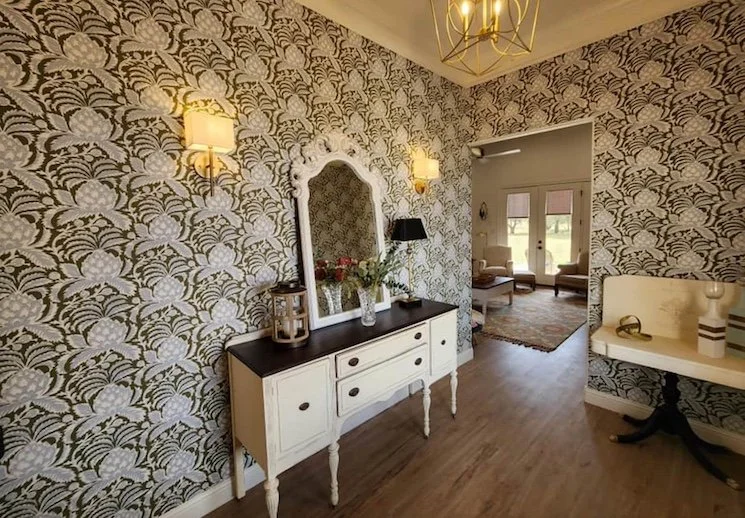 Entryway with floral wallpaper, a white console table with a mirror above it, a flower arrangement, two wall-mounted lights, and a glimpse into a sitting room with chairs and windows.