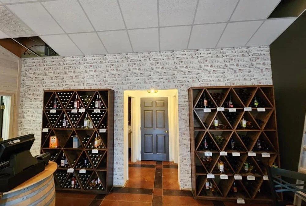Wine store with two large wooden wine racks filled with bottles, a doorway in the middle, and a cash register on a wooden counter on the left.