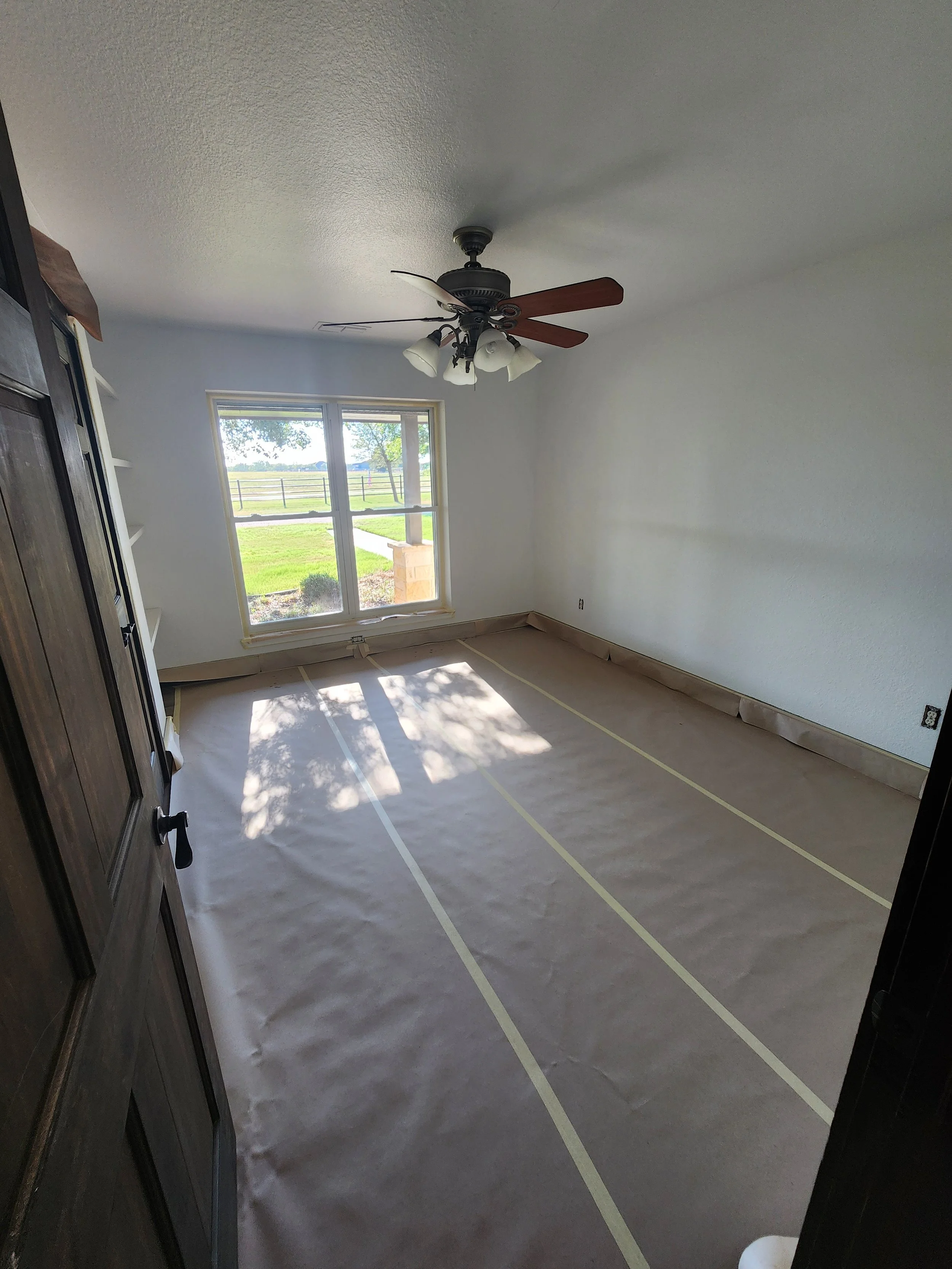 Empty room with white walls, window showing outdoor greenery, beige tape lines on the floor, ceiling fan with lights, and a dark wooden door.
