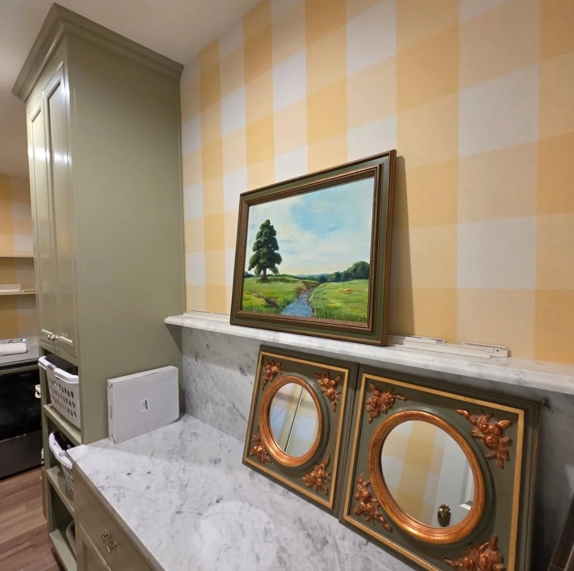 A framed landscape painting of a green field, river, and trees leaning against a checkered yellow and white wall, with two small decorative mirrors with gold floral accents on a marble countertop.