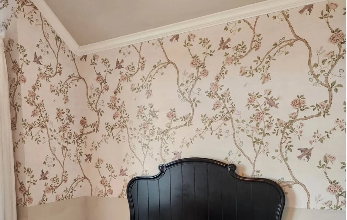 A bedroom corner with floral wallpaper featuring pink flowers, greenery, and birds, and a black headboard.