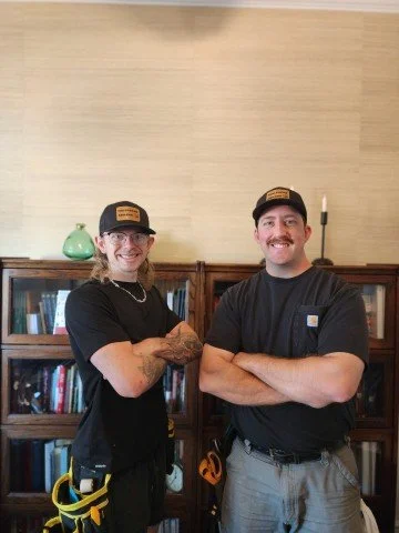 Two smiling men in black shirts and hats stand with arms crossed in front of bookshelves in a home setting. Residential wallpaper installation