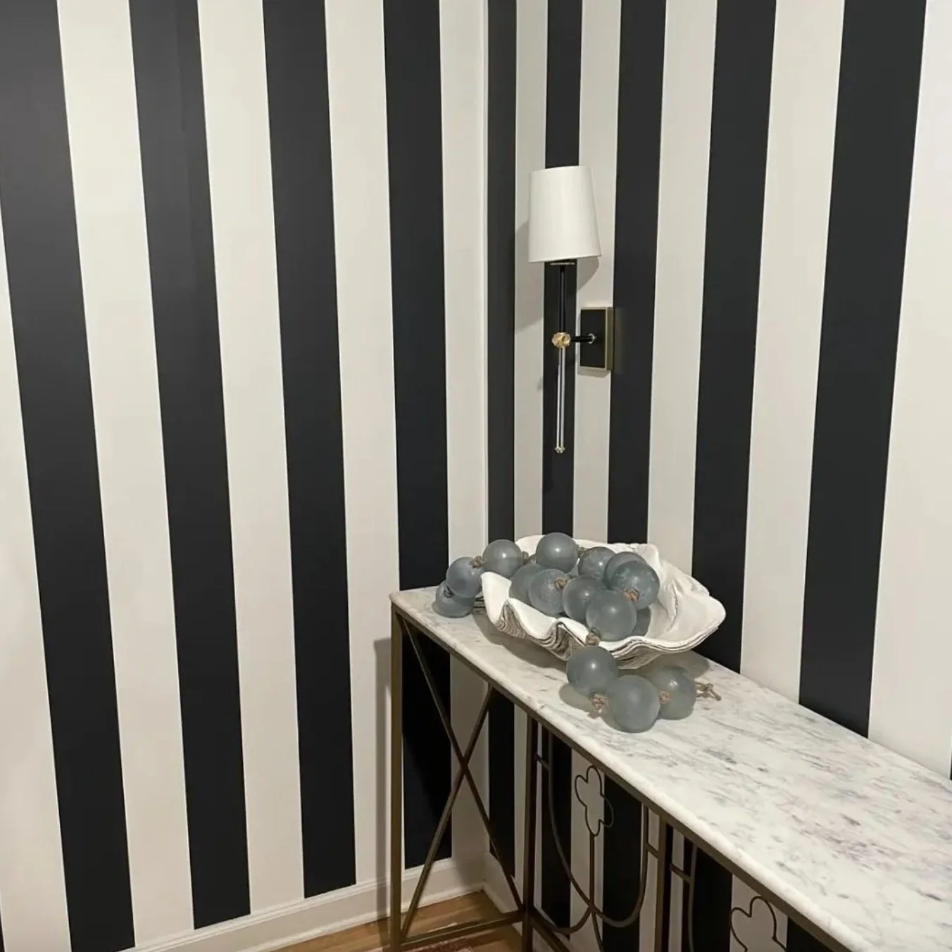 A corner of a room with black and white vertical striped wallpaper, a wall-mounted lamp, and a marble-top console table holding a white bowl with gray decorative spheres.