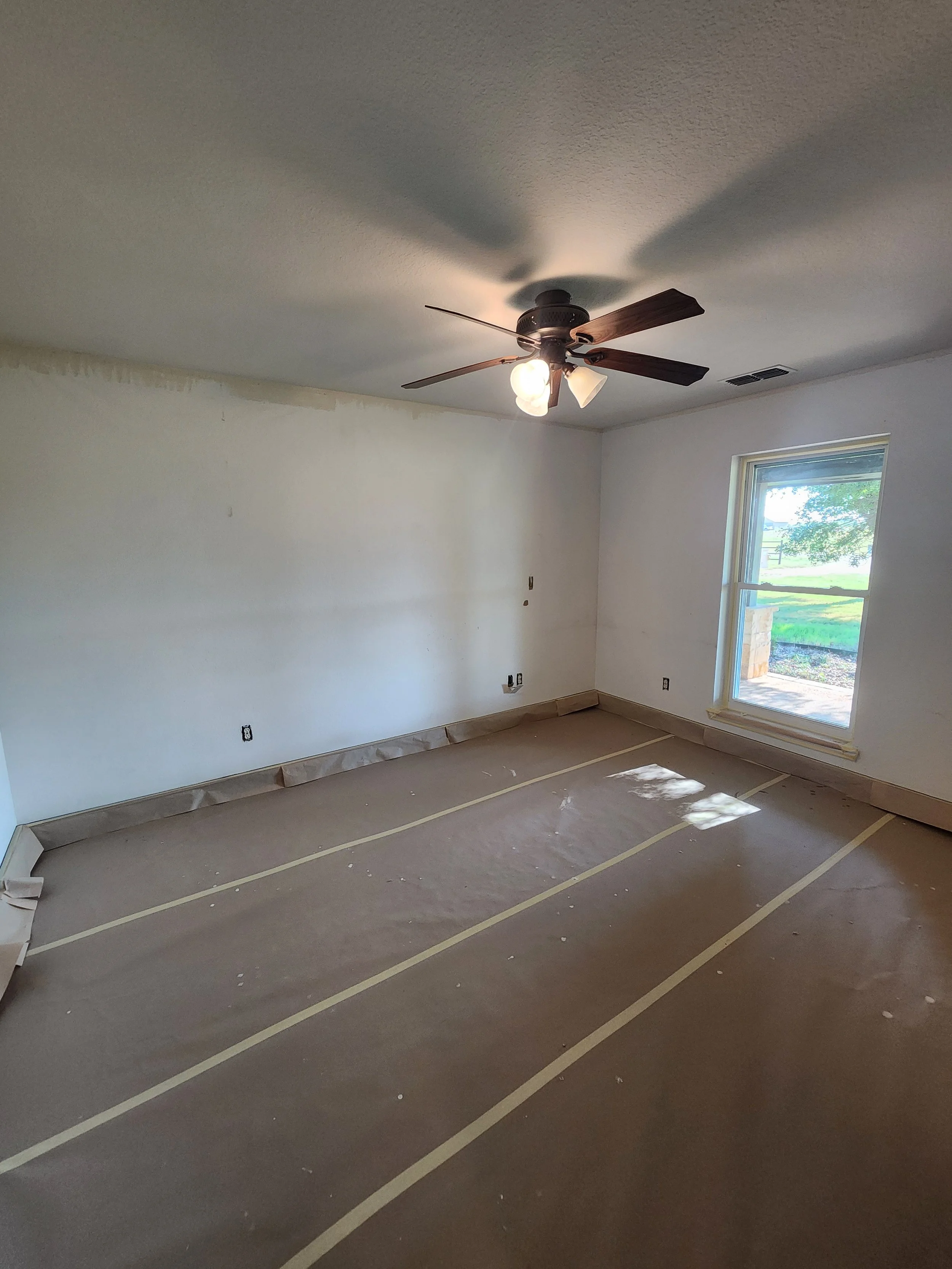 Empty room in a house under renovation with taped floor, white walls, a window on the right, and a ceiling fan with lights.