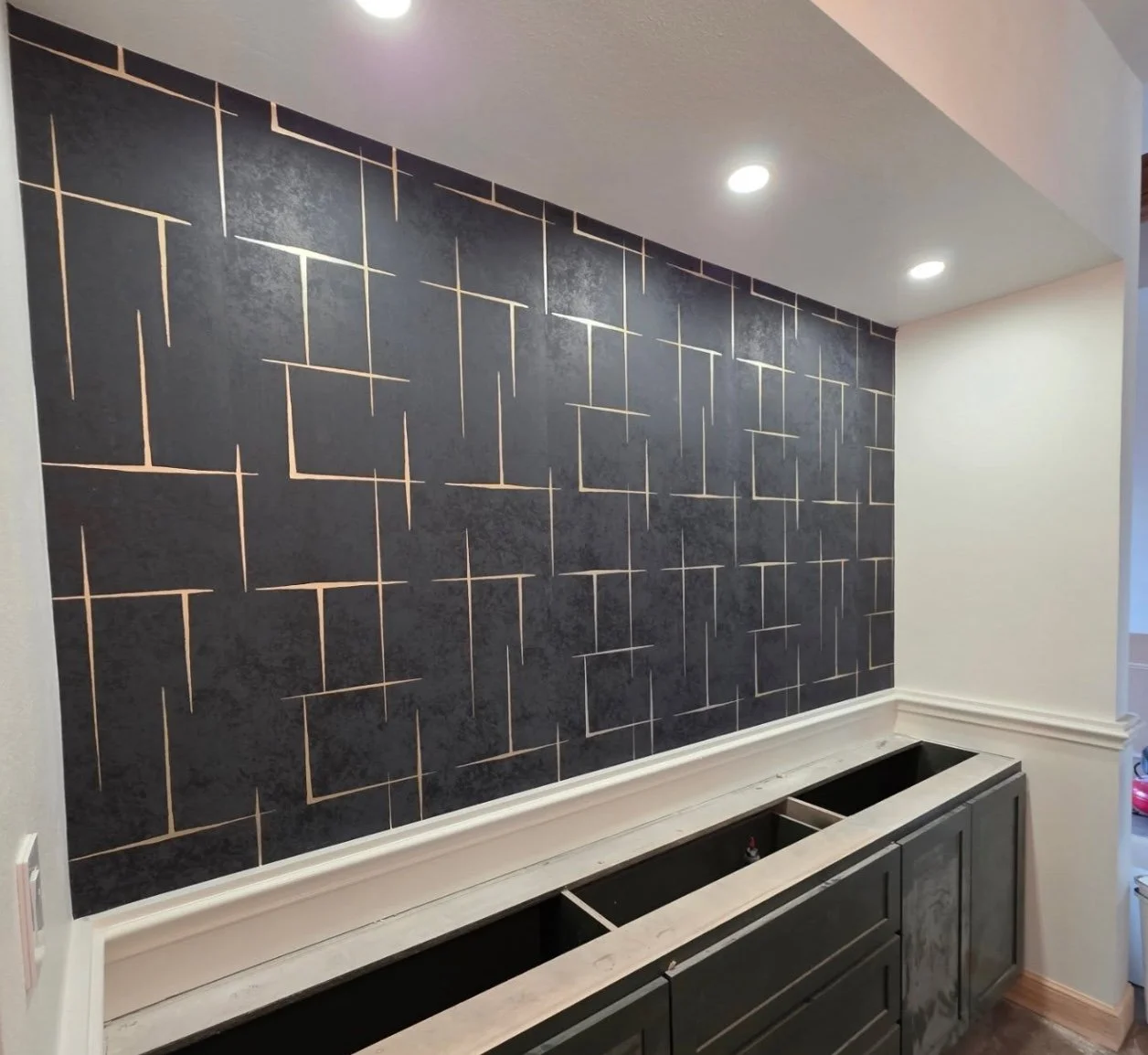 Interior wall with black textured tiles and geometric gold lines, with ceiling lights and dark cabinetry below.