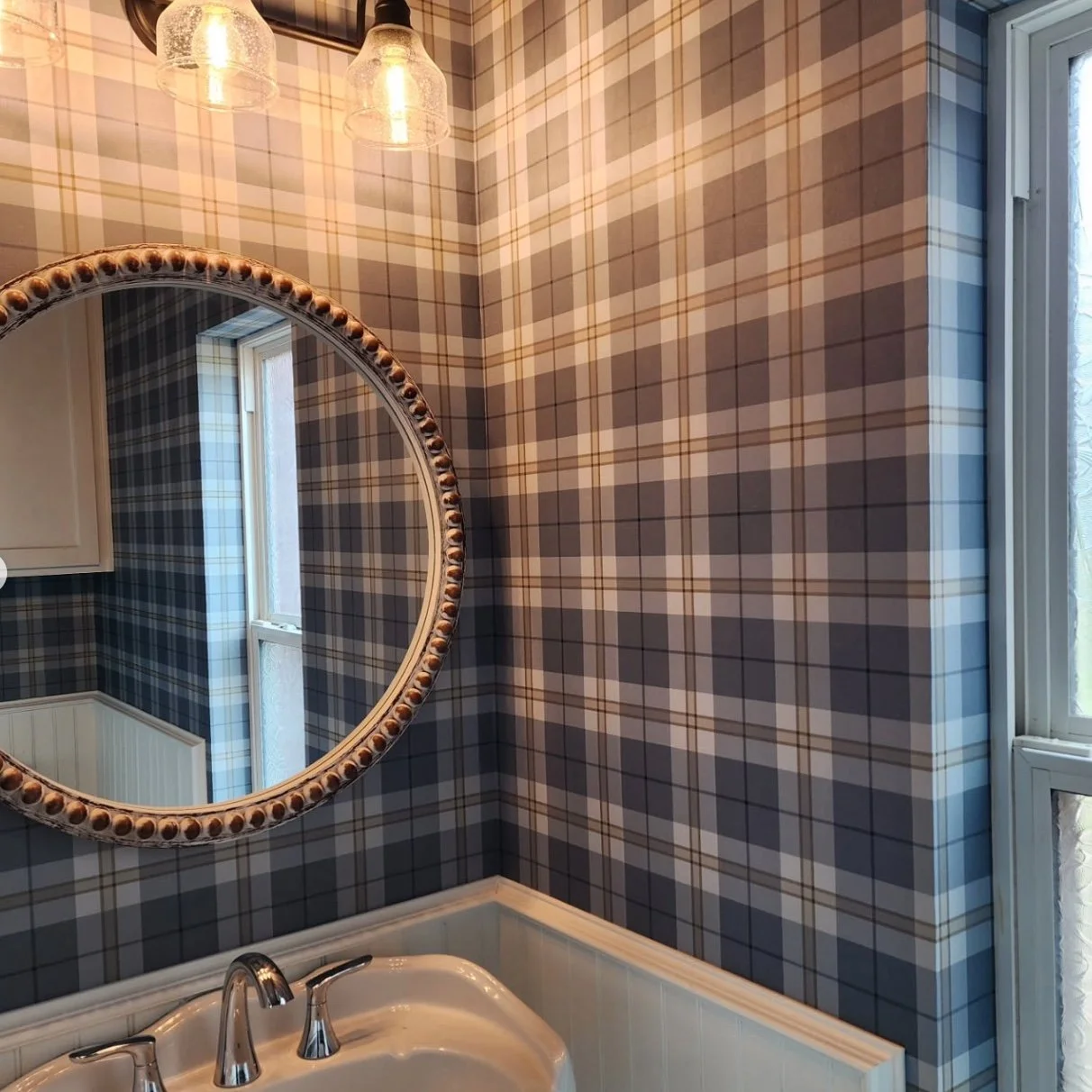 Bathroom with blue and white plaid wallpaper, a round mirror with decorative frame, a sink with silver faucet, and a window letting in natural light.