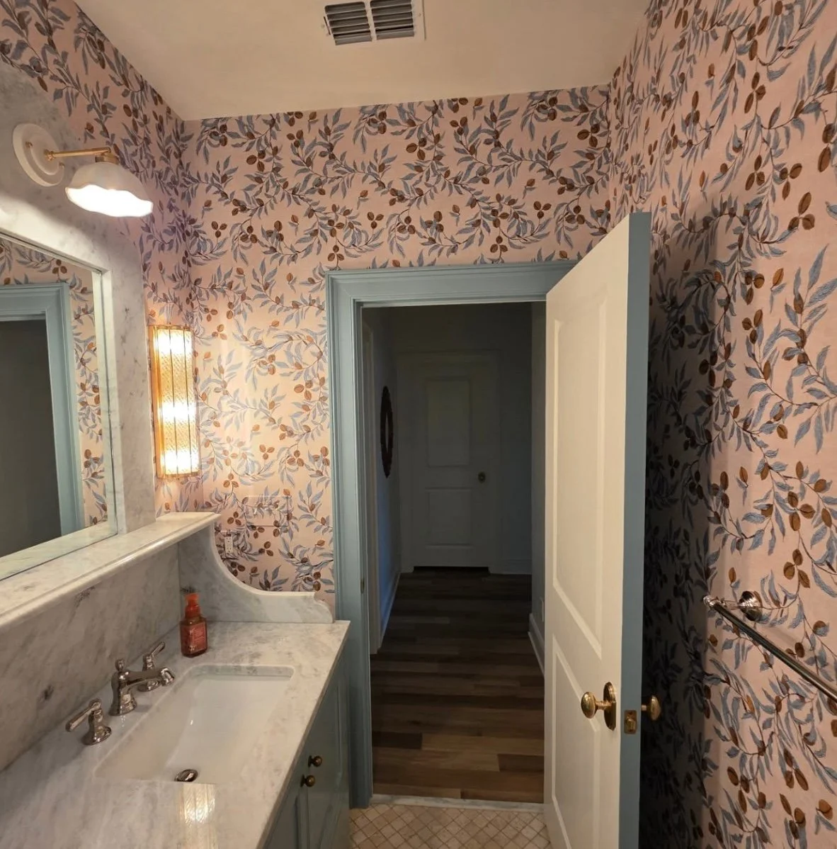 Bathroom with floral wallpaper, marble countertop with sink, mirror, wall-mounted light fixture, soap dispenser, door open to hallway with wood flooring, partially visible towel rack, and ceiling vent.