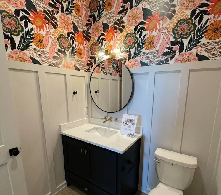 Bathroom with floral wallpaper on the upper half of the wall, white wainscoting on the lower half, a black vanity with a white countertop and sink, a round mirror above the sink, a wall-mounted light fixture above the mirror, and a toilet to the righ