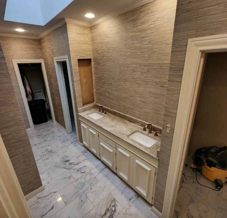 Bathroom with dual sinks, marble countertops, beige cabinets, textured beige walls, and overhead lighting.