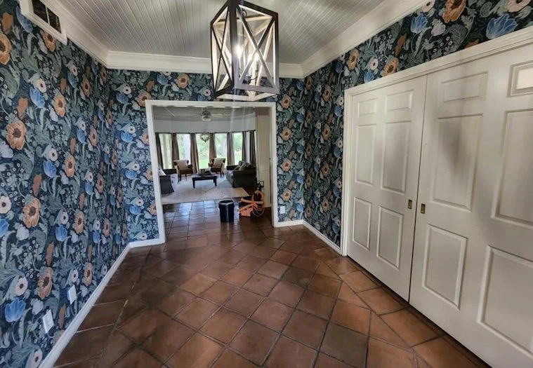 View of a room with floral wallpaper, tile flooring, and a ceiling light fixture, opening into a living room with chairs, a table, and large windows with curtains.