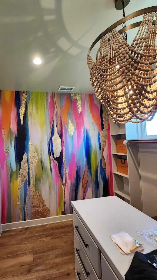 Colorful abstract wall mural with pink, blue, green, and gold accents in a modern room with a beaded chandelier and white cabinetry.
