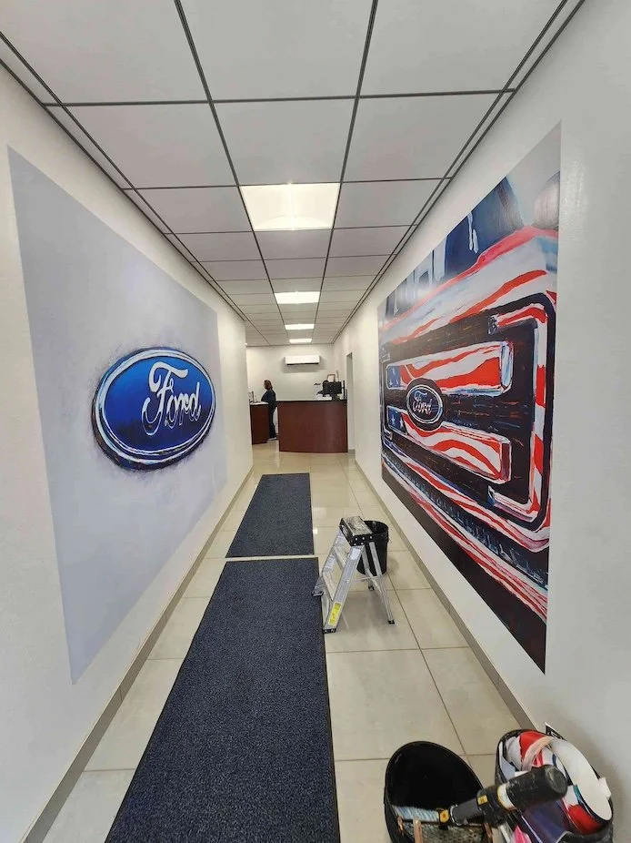 Interior of a Ford dealership showroom or service area with a Ford logo on the wall and a large Ford-themed artwork on the opposite wall. A small step ladder is placed on a dark carpet runner on the tiled floor, with a helmet or hard hat nearby.