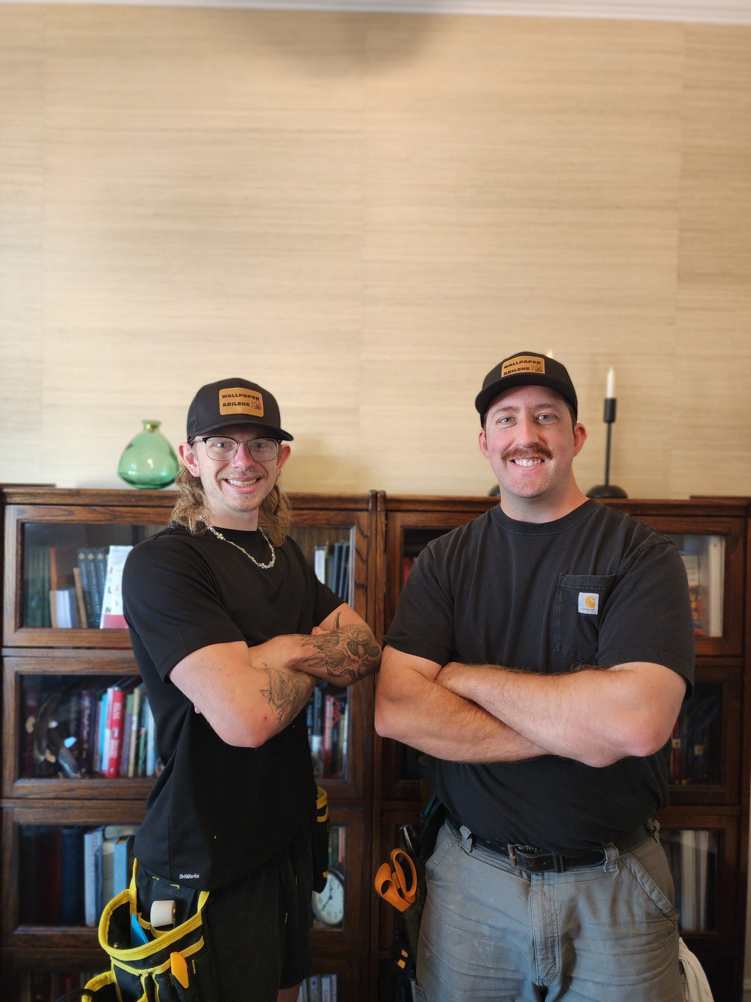 Two smiling men in black shirts and hats stand with arms crossed in front of bookshelves in a home setting.