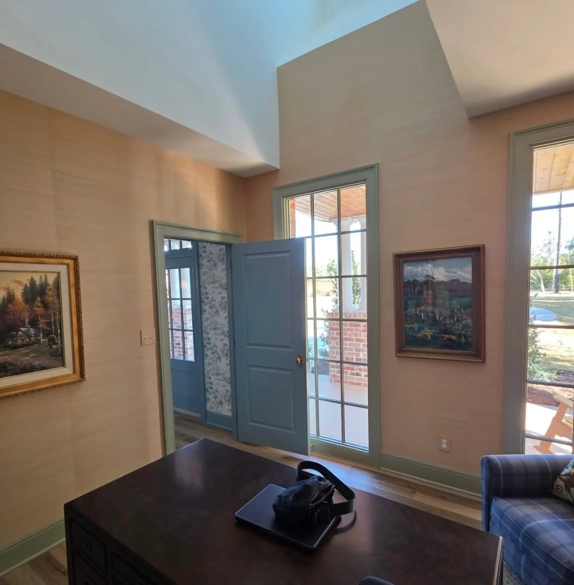 Interior of a living room with beige walls, large windows, and a door leading outside. There are framed landscape paintings on the walls, a dark wooden table with a backpack, and a blue checkered armchair. Light is coming through the windows.
