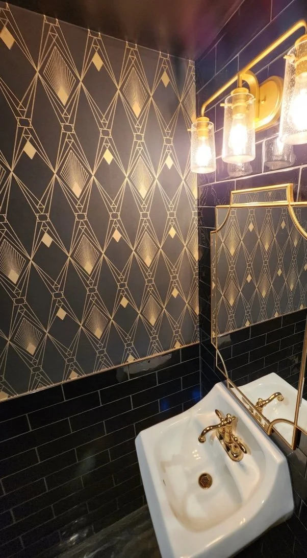 Elegant bathroom with geometric gold and black wallpaper, gold fixtures, glass light bulbs, and a white sink below a mirror.