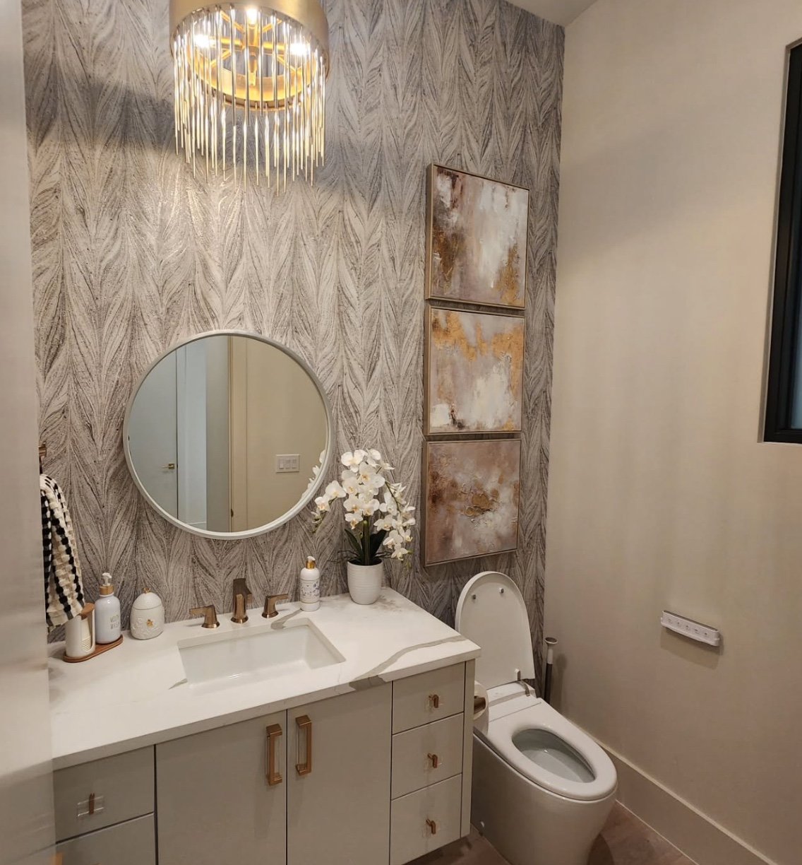 Modern bathroom with a white vanity, round mirror, decorative paintings, and a toilet, with a chandelier and patterned wallpaper.