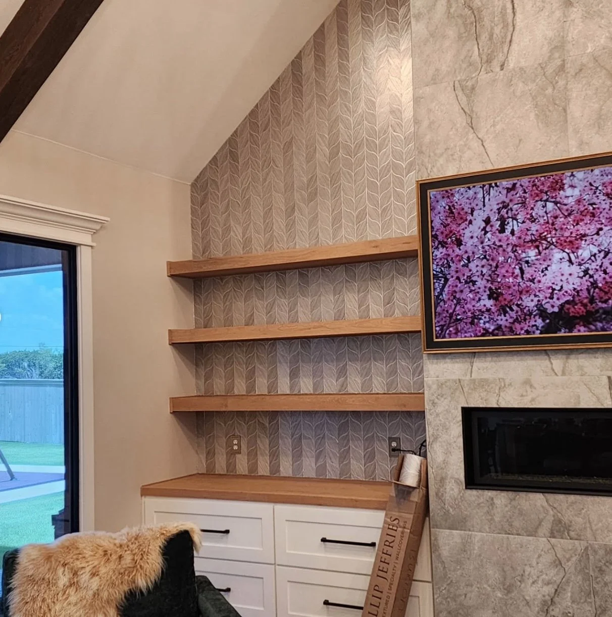 Living room with a built-in wooden shelf unit, a wall-mounted television displaying pink cherry blossoms, a fireplace, a sliding glass door, and a piece of wall art leaning against the wall.