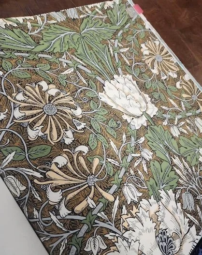 Close-up of a fabric with a floral pattern featuring white, beige, and green flowers and leaves on a brown background.