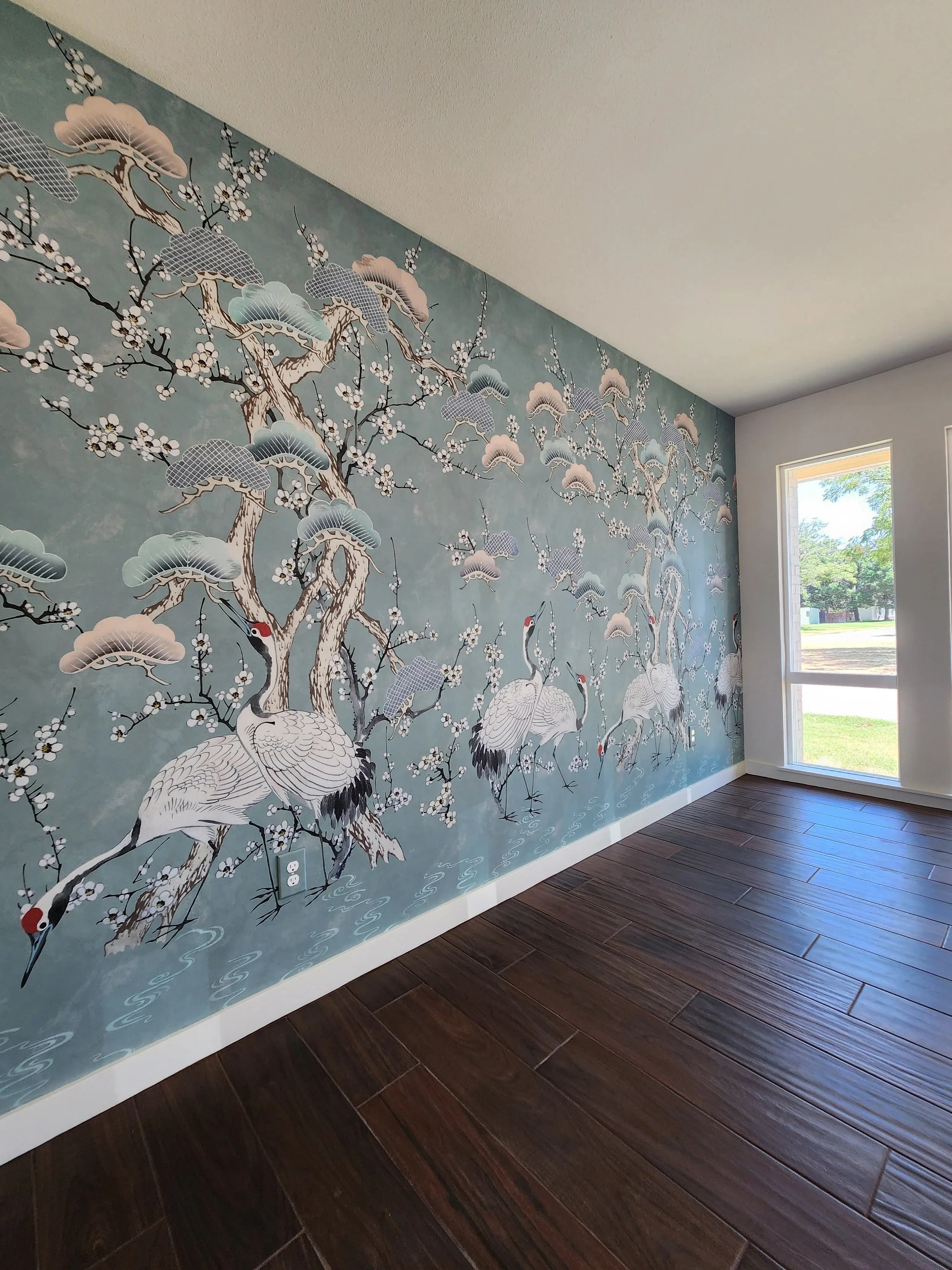 Interior of a room with a large mural of cranes, cherry blossoms, clouds, and water in Asian art style on the wall, hardwood flooring, and windows letting in natural light.