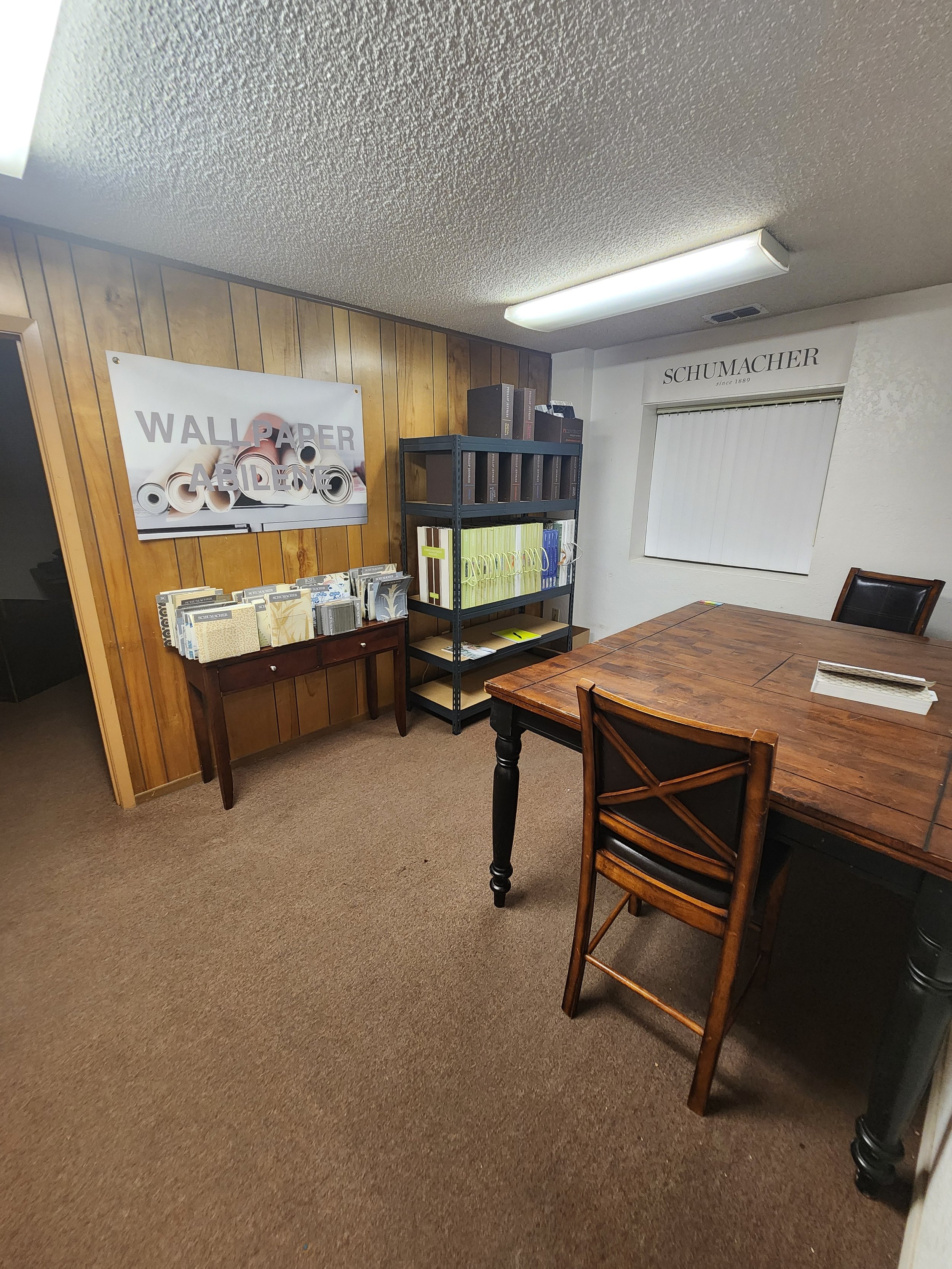 An office with wood-paneled wall, a wooden table with chairs, a shelving unit with folders, a small table with brochures, and a wall sign that says "SCHUMACHER."