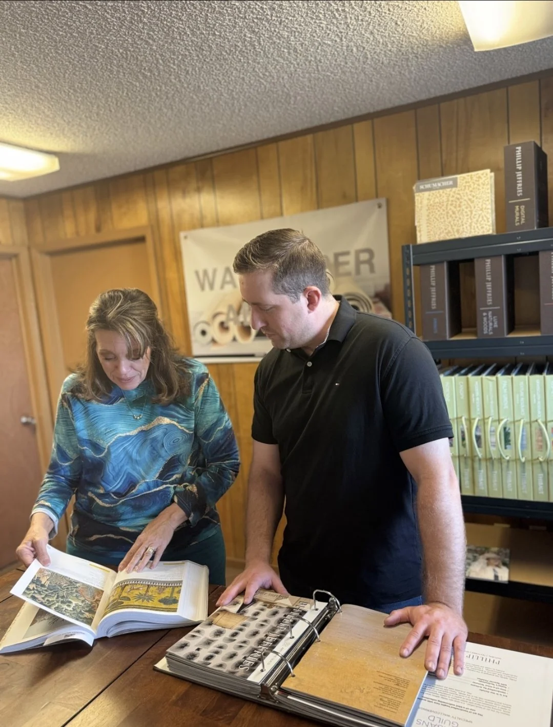 Two people, a woman and a man, are looking at a large photo album or book on a wooden table, examining images inside. The setting appears to be an office or a library with wood-paneled walls and shelves filled with boxes and documents in the backgrou