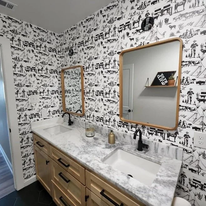 Double bathroom vanity with white and gray marble countertop, wooden cabinets, two black faucets, and two framed mirrors on patterned black and white wallpaper wall.