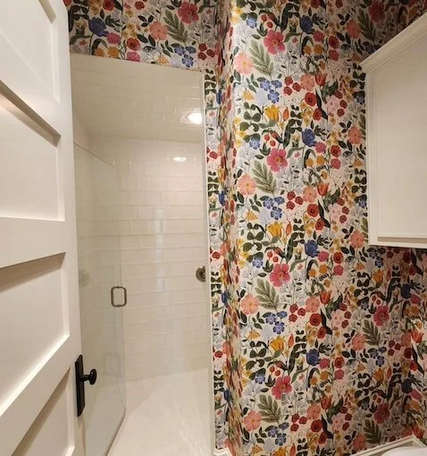 A bathroom with a floral wallpaper pattern and a glass shower door. Part of a white cabinet and door with black handle are visible.