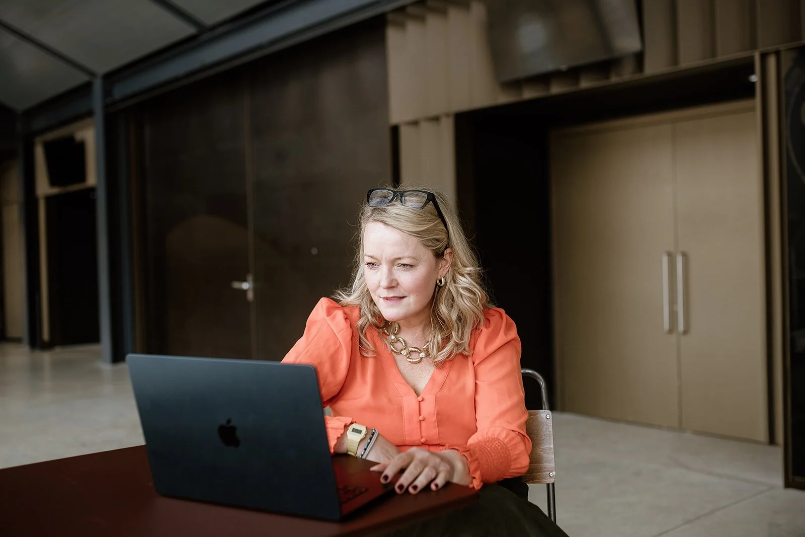 Lucy Timmons, Coach, A woman with blonde hair, wearing a coral blouse, glasses on her head, and a gold necklace, working on a black laptop at a table in an indoor setting.