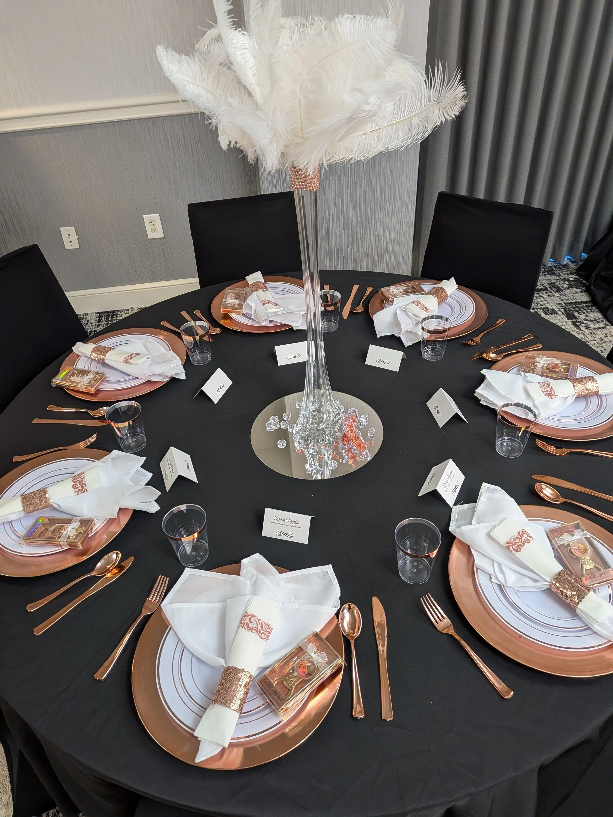 Round table set for a formal event with a black tablecloth, six place settings, and a tall feather centerpiece. Each setting includes a white napkin with a decorative band, a clear favor box, and a rose gold charger plate. There are black chairs around the table, grey walls, and a mirrored base under the centerpiece.