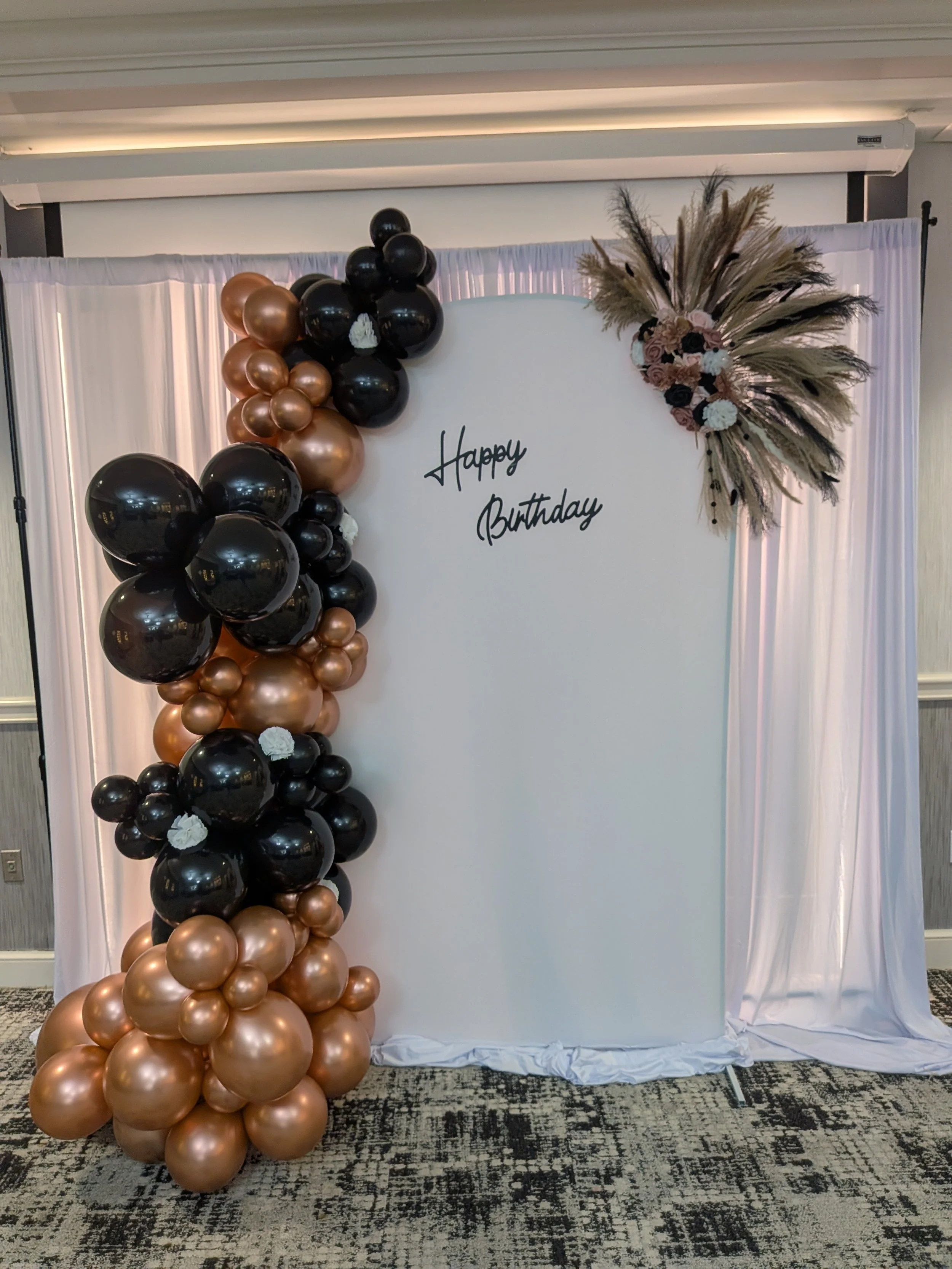 Balloon backdrop with black, gold, and bronze balloons arranged vertically, floral decoration with dried pampas grass and roses on the top right, white backdrop with a 'Happy Birthday' sign in black script.