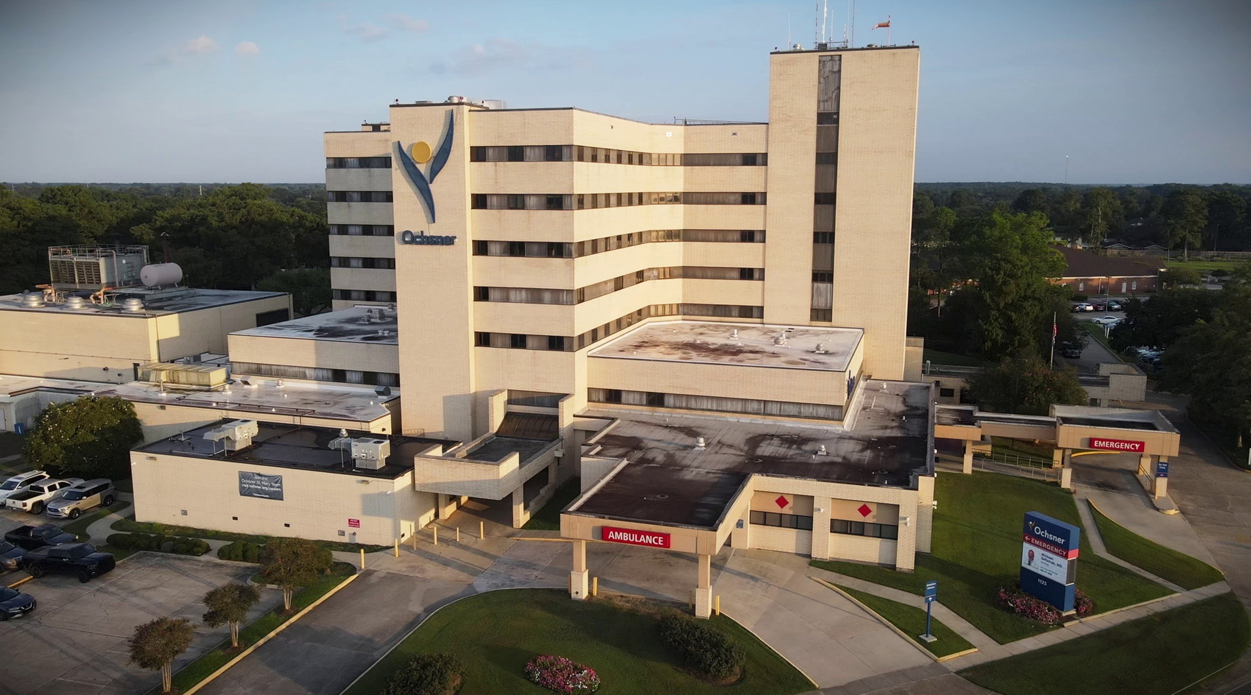 Aerial view of Ochsner Medical Center with multiple buildings, parking lot, and entrance signs for emergency and ambulance services.