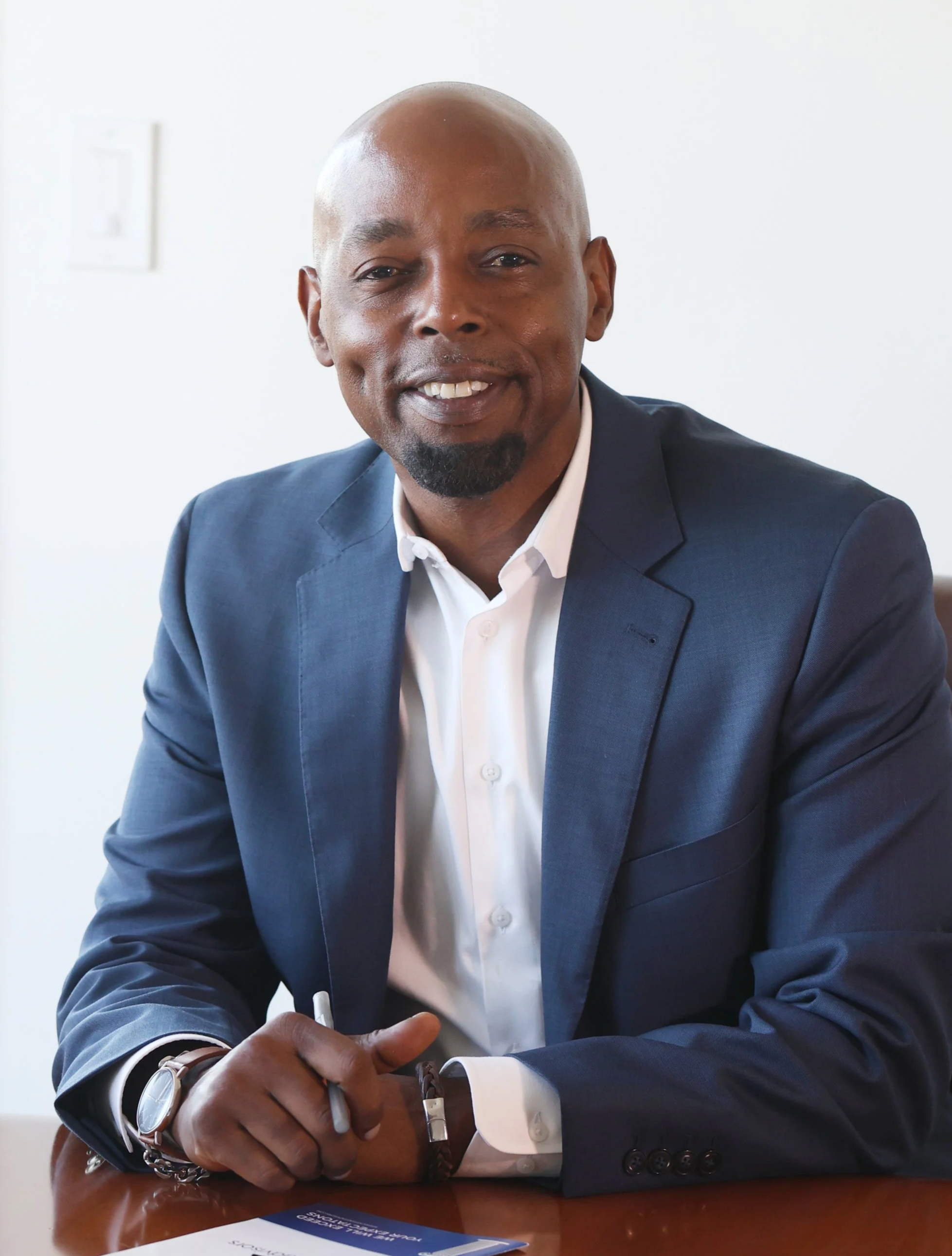 Man in navy suit and white shirt sitting at a table, smiling, holding a pen, with a document in front of him.