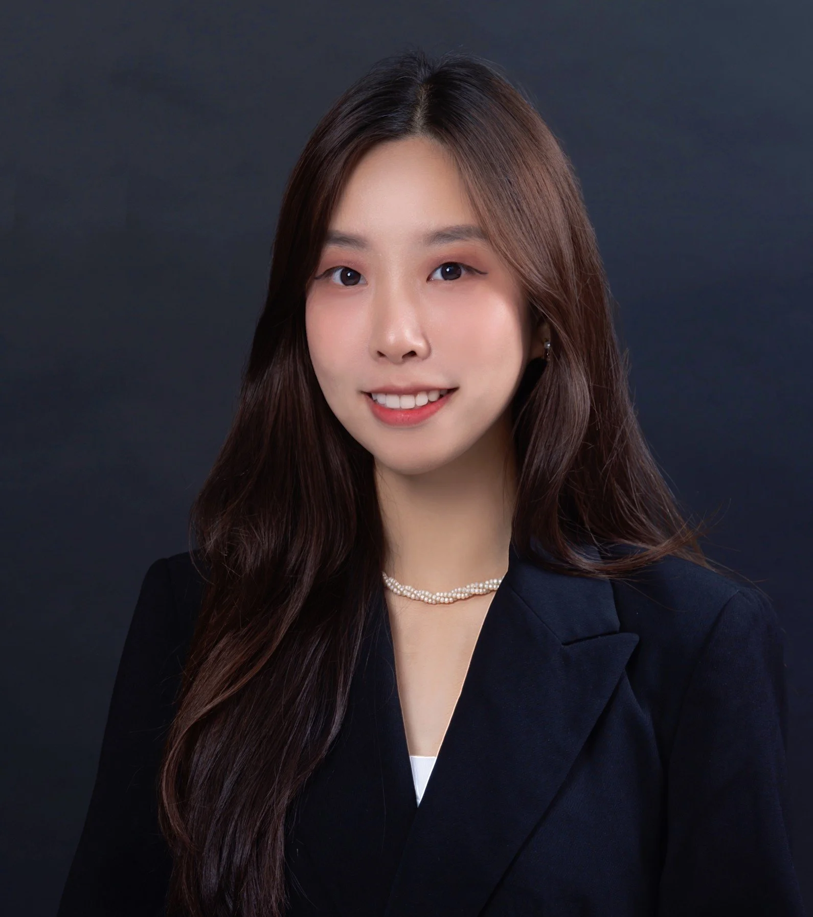 A young woman with long wavy brown hair winking and smiling at the camera, wearing a dark blazer, pearl necklace, and earrings against a dark background.