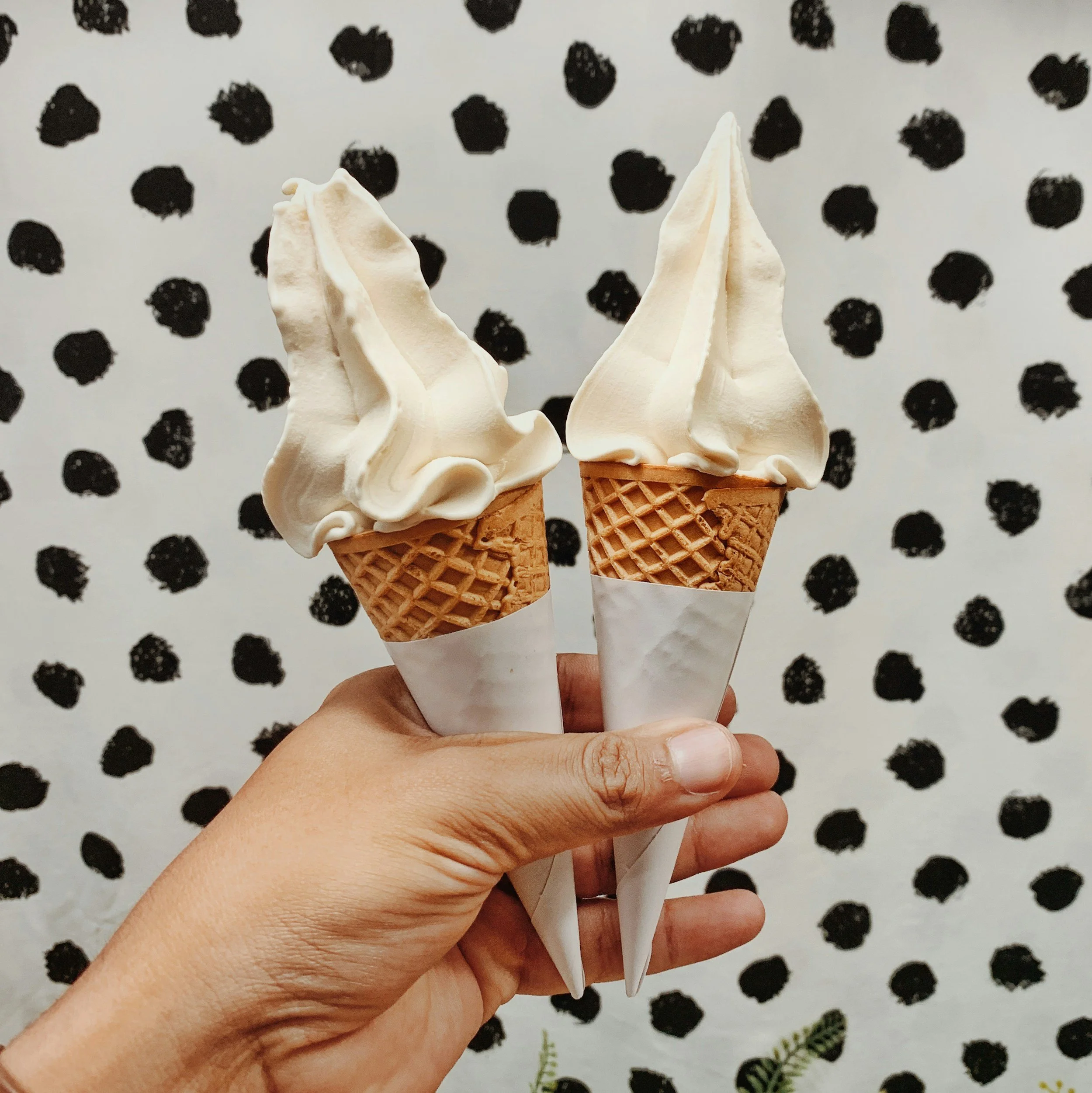 zwei Eiscreme in Waffelhörnchen, gehalten in der Hand, vor einem Hintergrund mit schwarzen Tupfen auf weißem Untergrund