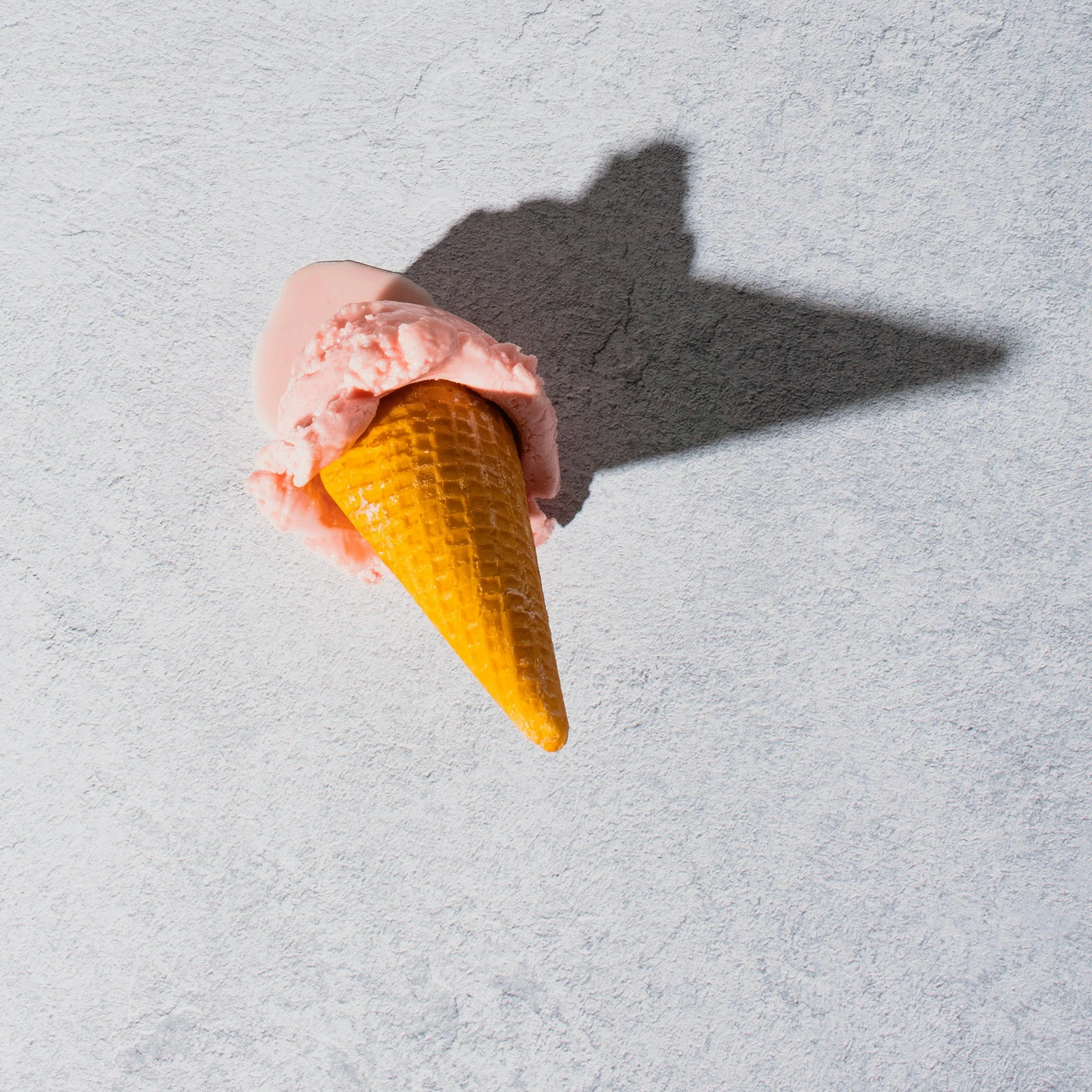 Ein Eis in Waffel mit einer Kugel Vanille und einer Kugel Erdbeere auf einer weißen Oberflächenstruktur, Schatten ist sichtbar.