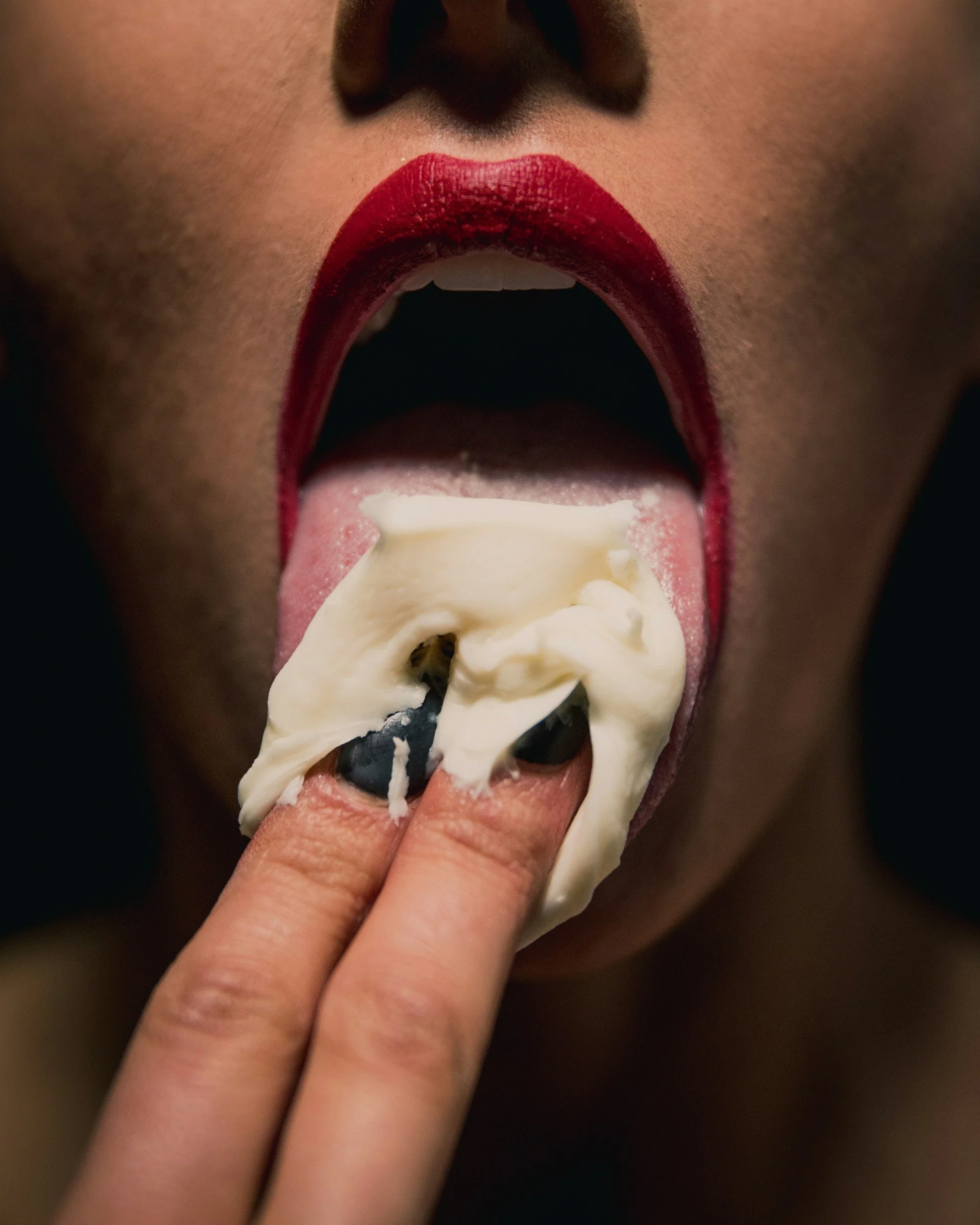 Nahaufnahme eines Mundes mit rotem Lippenstift, Zunge und Fingern, die auf der Zunge ruhen, sichtbar. Das Bild zeigt die Nasenöffnung und einen Teil der Wangen.