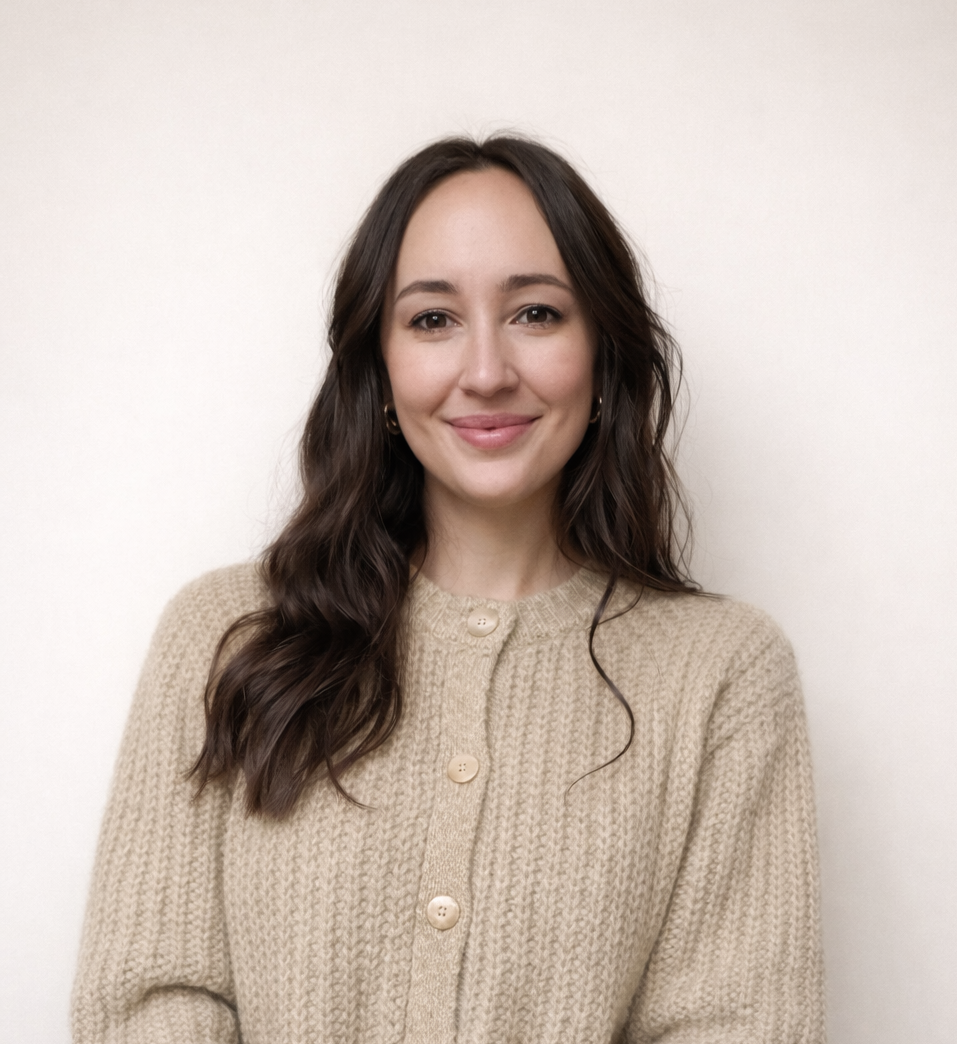 A woman with long, wavy brown hair and light makeup smiling, wearing a beige knit cardigan with buttons, standing against a plain white wall.