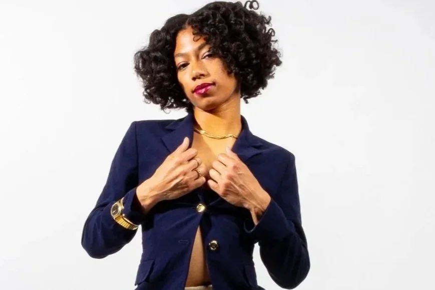 A woman with dark curly hair posing against a white background, wearing a dark blue blazer with gold buttons and gold jewelry.